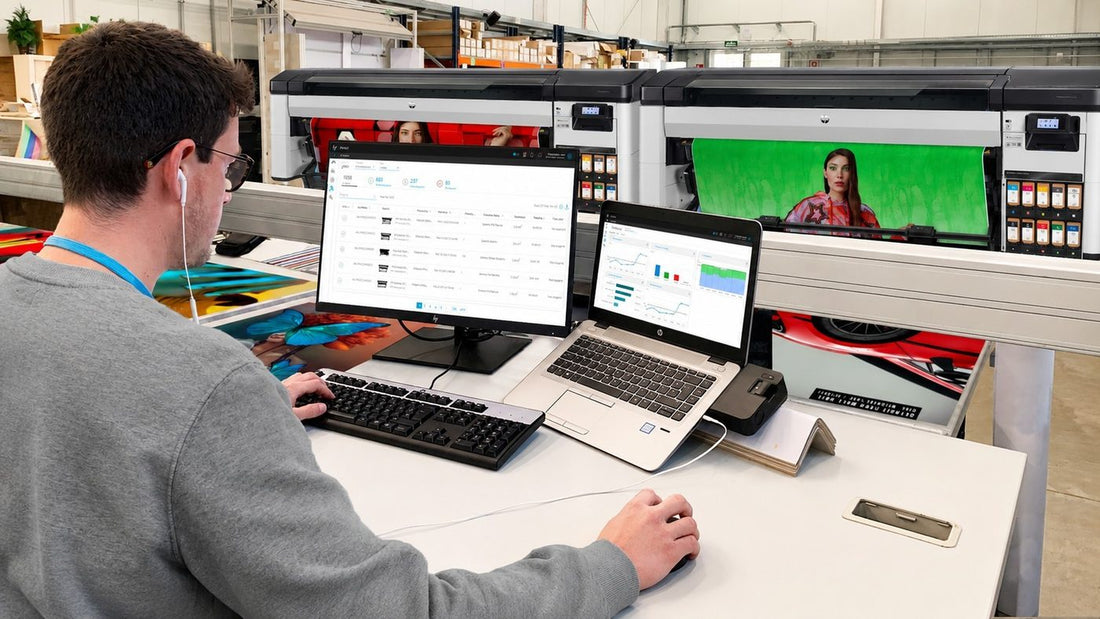 Employee working at a computer in a print production workspace with large-format printers, showing workplace technology that supports productivity.