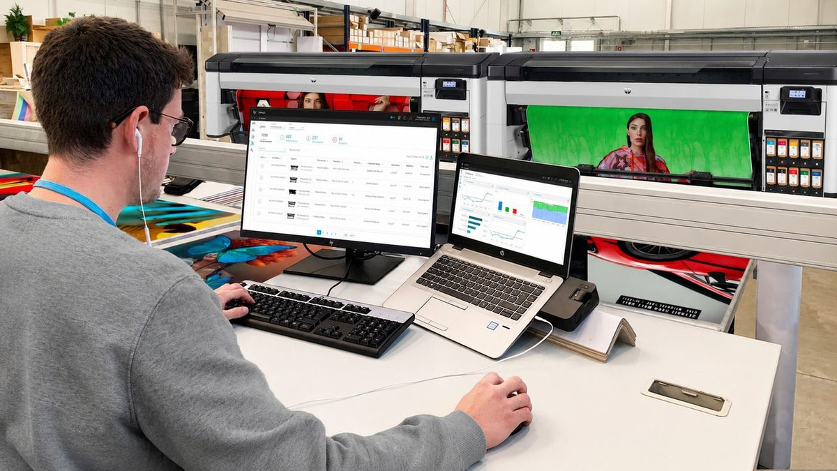 Employee working at a computer in a print production workspace with large-format printers, showing workplace technology that supports productivity.